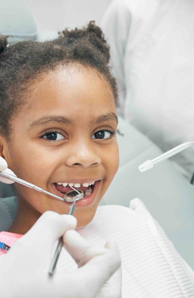 Happy afro american girl sitting in stomatologist chair with open mouth while professional dentist doing regular check up of teeth using dental probe and mirror. Female nurse assisting.